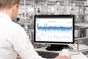 person sitting at a desk looking at a monitor with a graph, manufacturing environment in the background